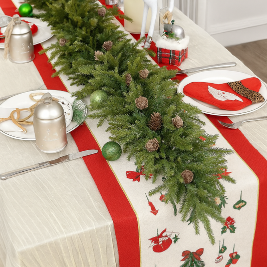 Christmas-themed table setting with greenery, ornaments, and red ribbon on a white tablecloth.